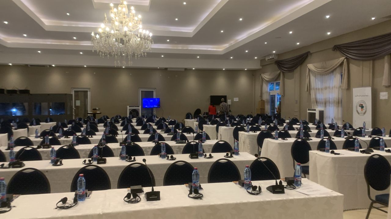 Man sitting at a white table with arms crossed, smartphone placed on the table, bright window in the background.
