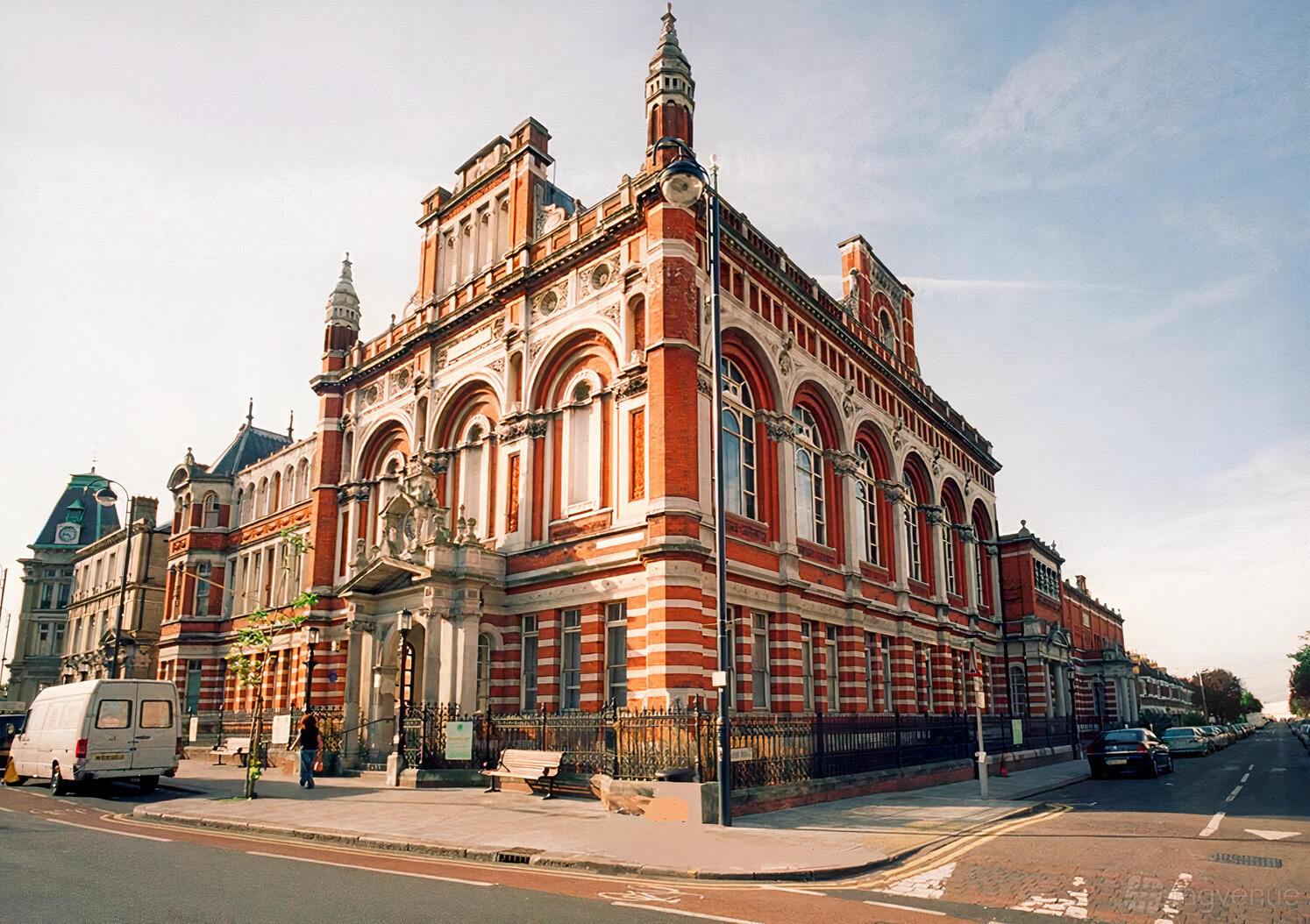 Historic red brick building with ornate architectural details on a street corner, featuring arched windows and decorative turrets under a clear blue sky.