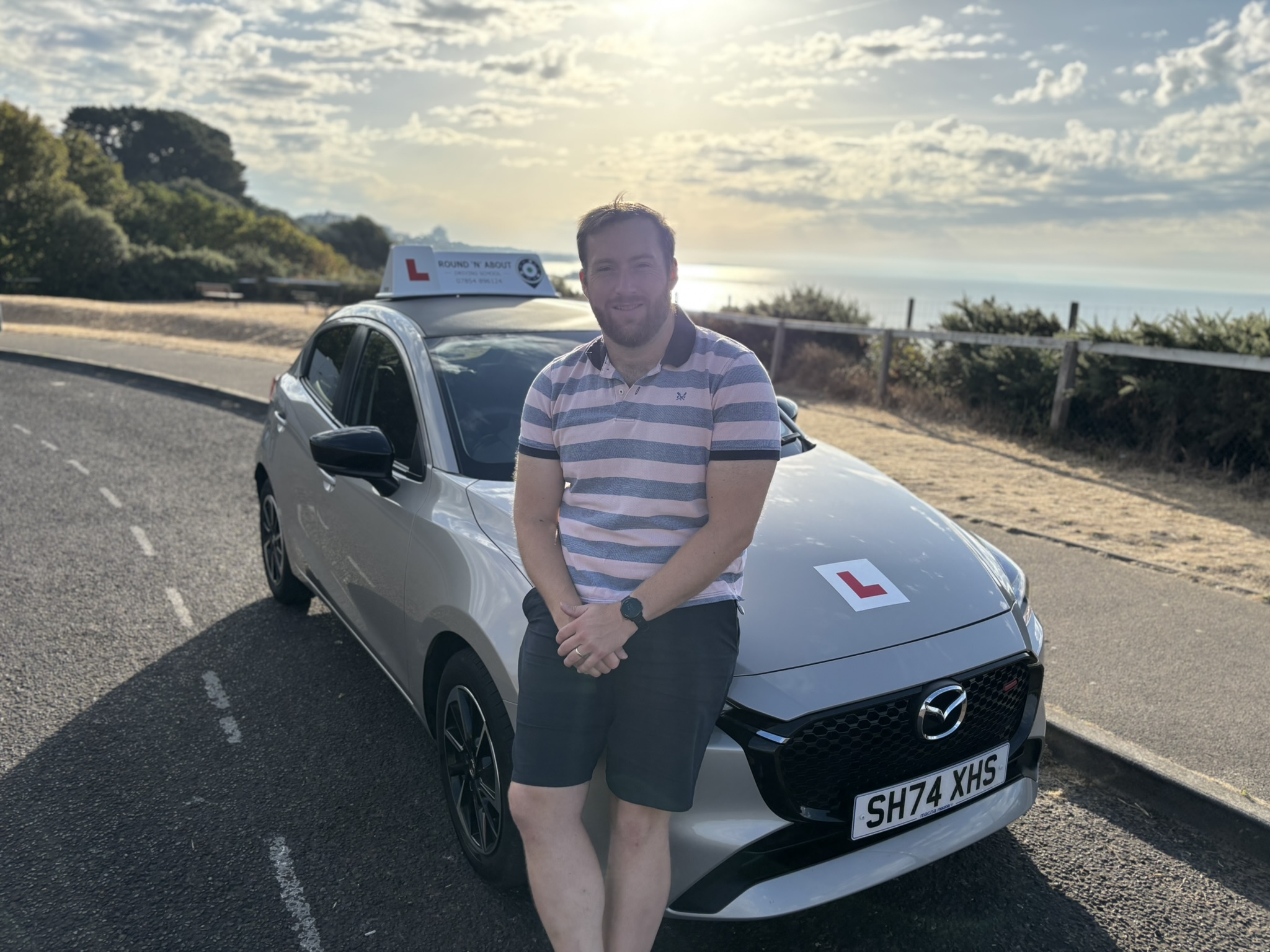 Silver Mazda driving school car parked by coastal road with ocean view and blue sky