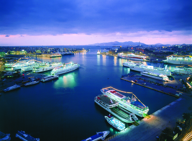 Aerial view of a vibrant harbor at dusk with illuminated cruise ships and city lights reflecting on calm waters.