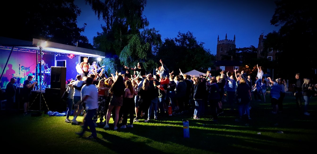 Outdoor music festival at night with a band performing on stage and a crowd dancing on the grass in front of a historic building.