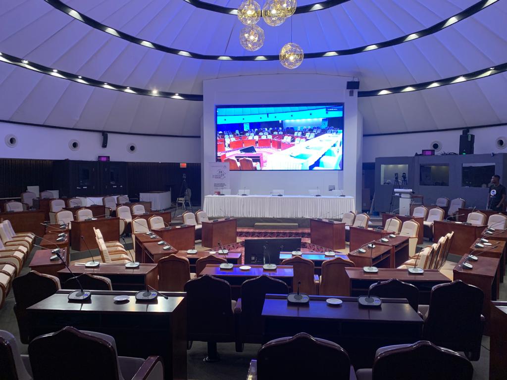 Person wearing a skeleton mask with a stethoscope, standing in front of digital screens displaying data, in a dimly lit room.