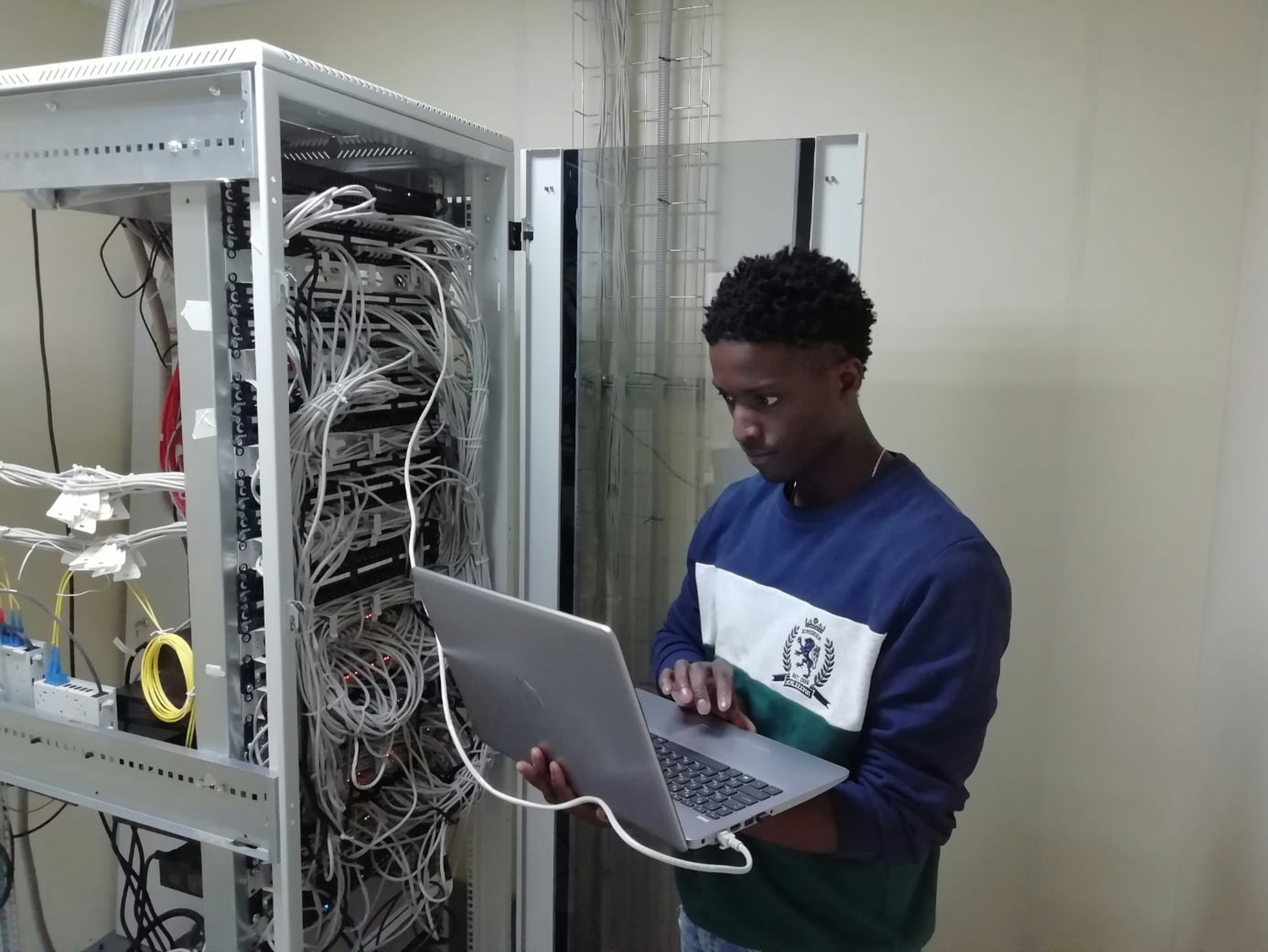 Person using a laptop near open server rack with numerous cables in a data center. Person using a laptop near open server rack with numerous cables in a data center.