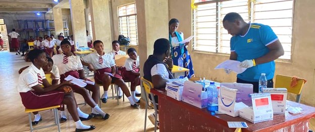 Students in uniforms sitting in line for a medical check-up while healthcare workers prepare documents and medical supplies on a desk in a classroom setting.