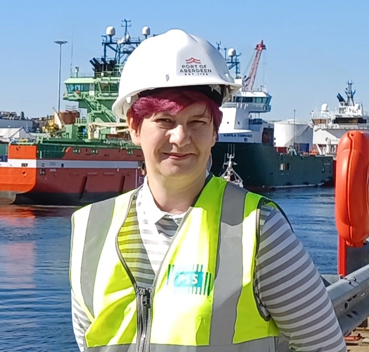 Debbie Cavaldoro chair of the NUJ's Standing Orders Committee wearing a hard hat and hi-viz vest standing in front of a commercial ship.
