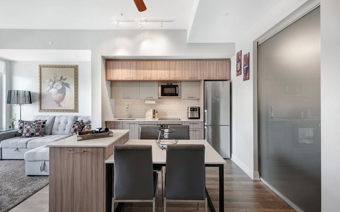 Modern open-plan kitchen and living area with wooden cabinets, stainless steel appliances, grey sofa, and stylish decor.