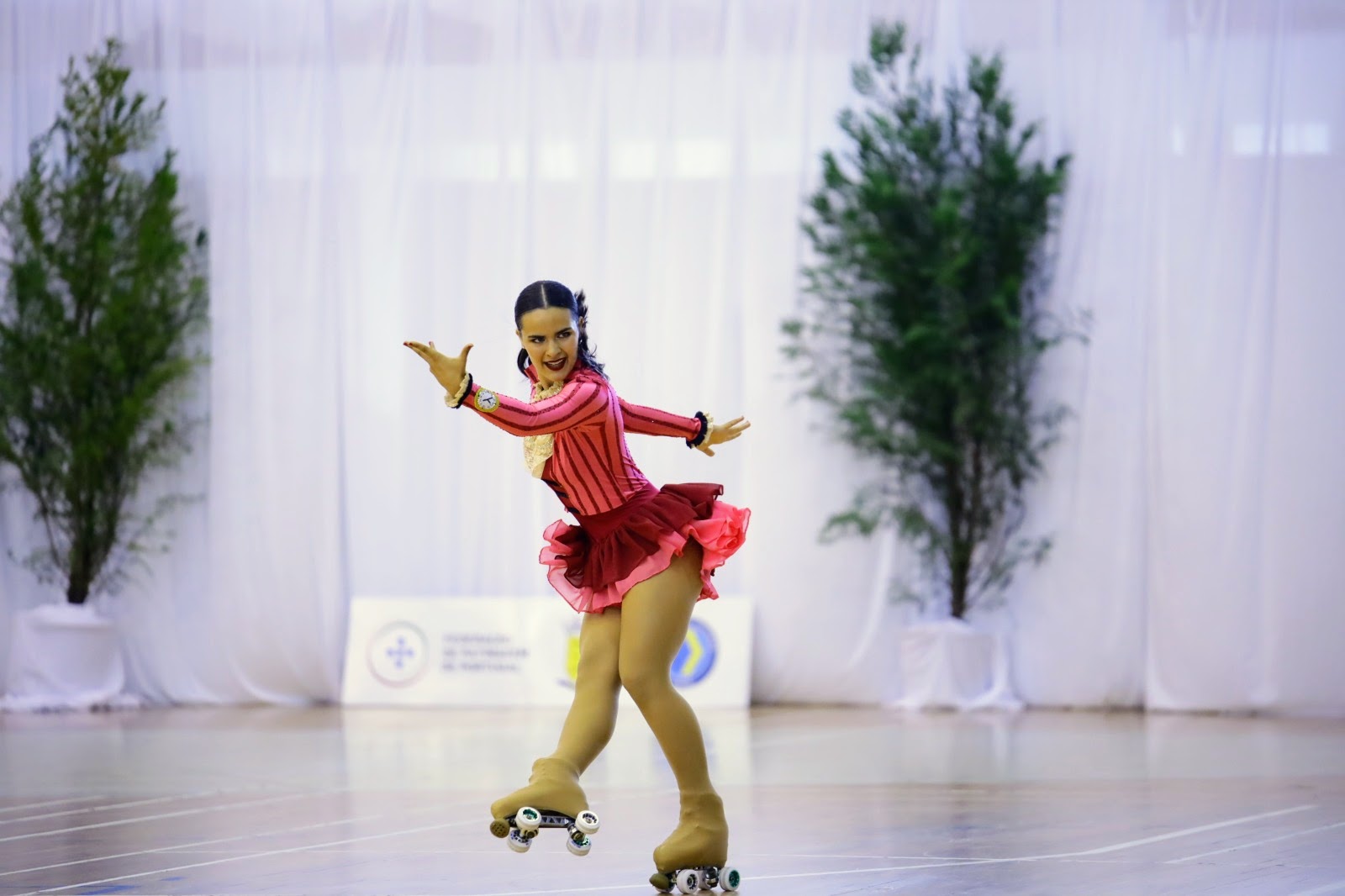 Patinadora artística fazendo uma apresentação em patins, com vestido rosa vibrante, sobre pista de madeira em ambiente decorado com plantas.