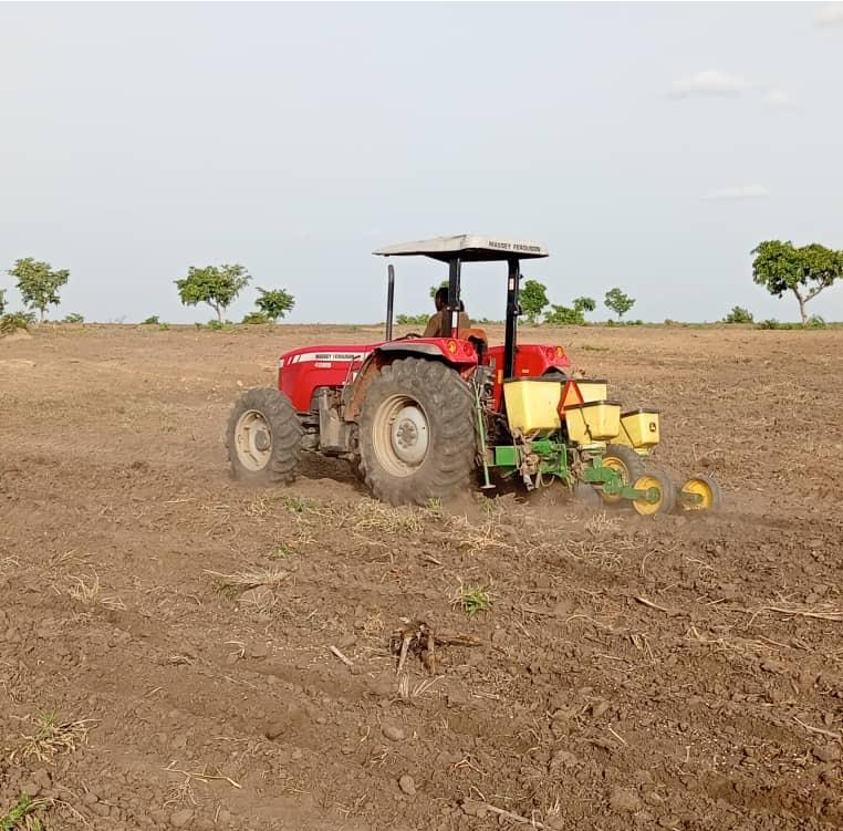 Agricultural workers inspecting crops in a farm field with tractors, trucks, and grain silos in the background