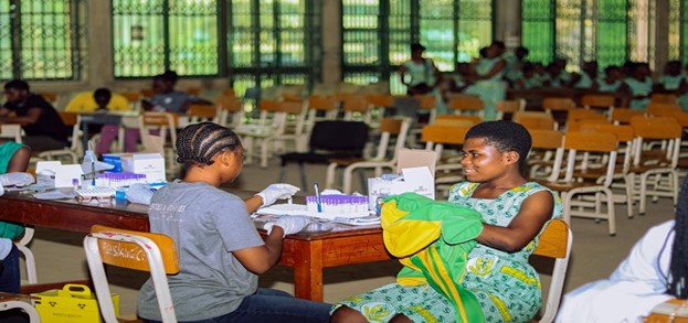 Health worker conducting a medical test for a woman in a clinic setting with various medical supplies on a table, surrounded by empty chairs.