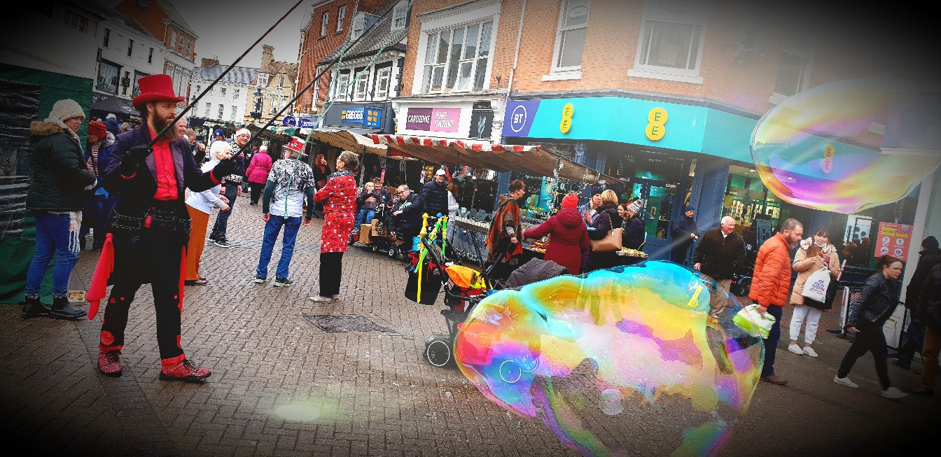Street performer creating large colorful bubbles in a busy marketplace with people shopping and observing.