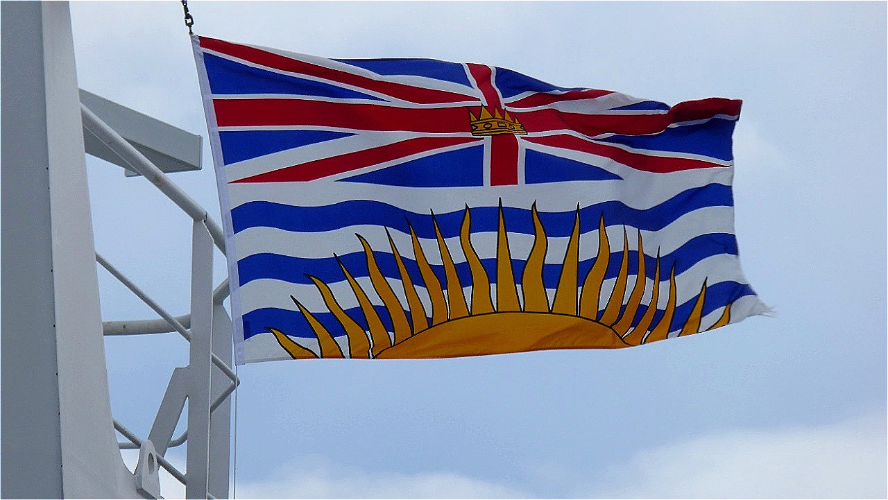 
Flagge von British Columbia:  oberes Drittel Union Flag Großbritanniens mit Krone; Mitte und unten: drei blaue Wellenbalken, die von der untergehenden Sonne überstrahlt werden.
