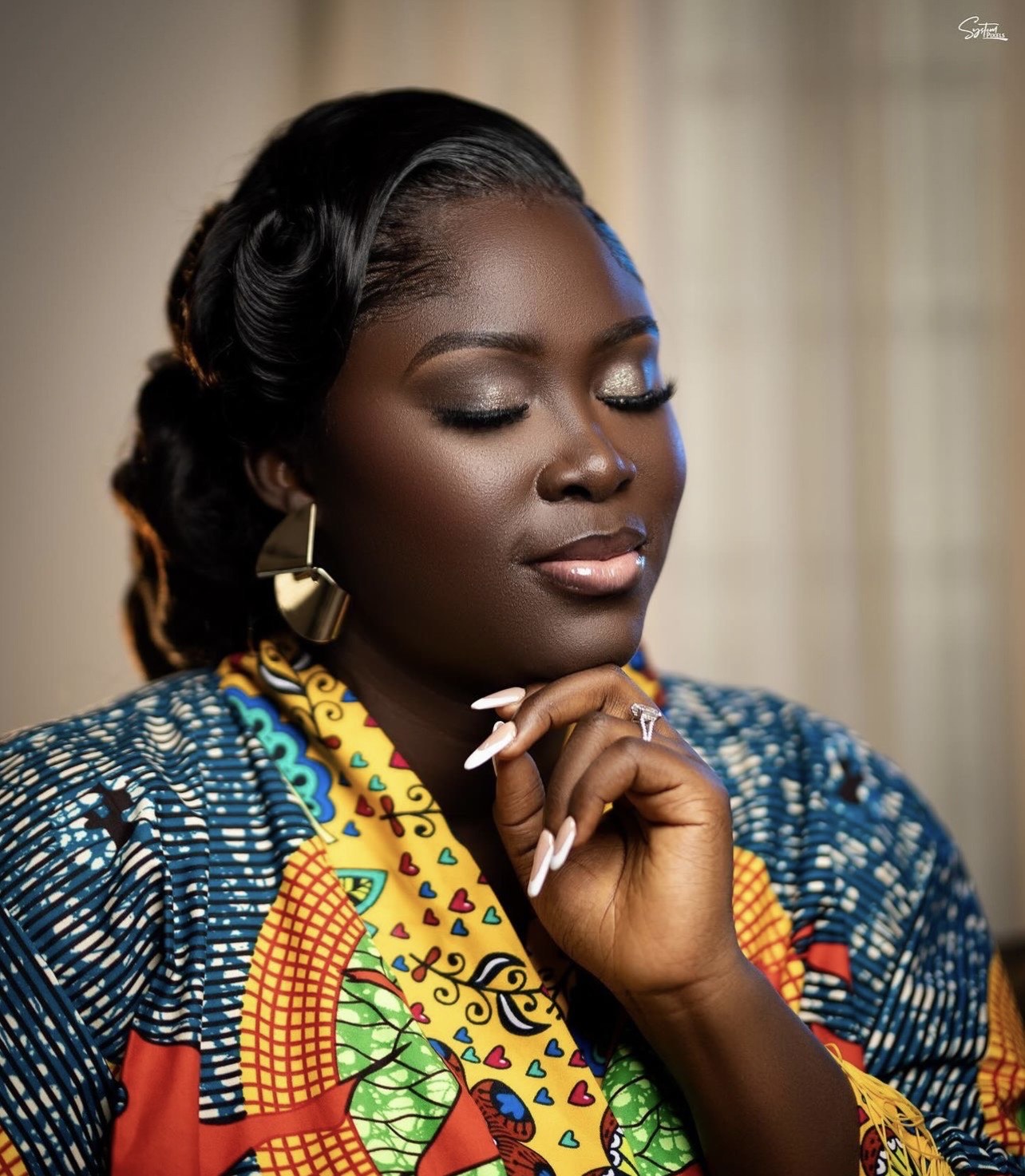 Portrait of a woman with shoulder-length blonde hair and a white dress in a sunlit room with plants in the background.