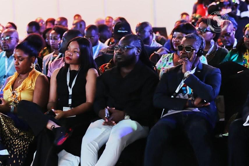 Young boy eagerly biting into a slice of watermelon while a baby observes, set against a brick wall background.