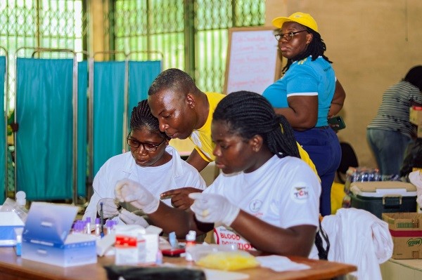 Medical professionals conducting blood tests at a health camp, with supplies and participants in the background.