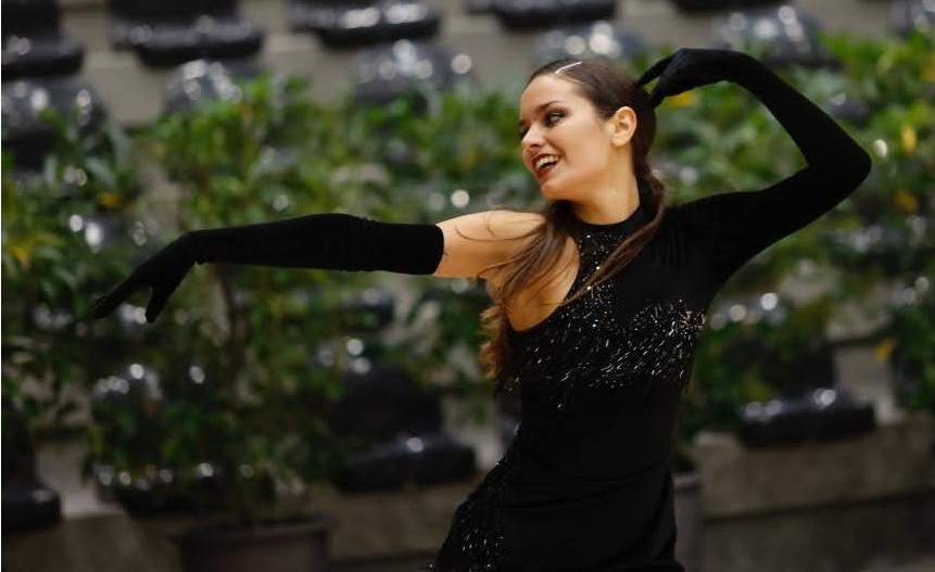 Dançarina com vestido preto brilhante e luvas longas, realizando uma pose expressiva em um ambiente interno com plantas ao fundo.
